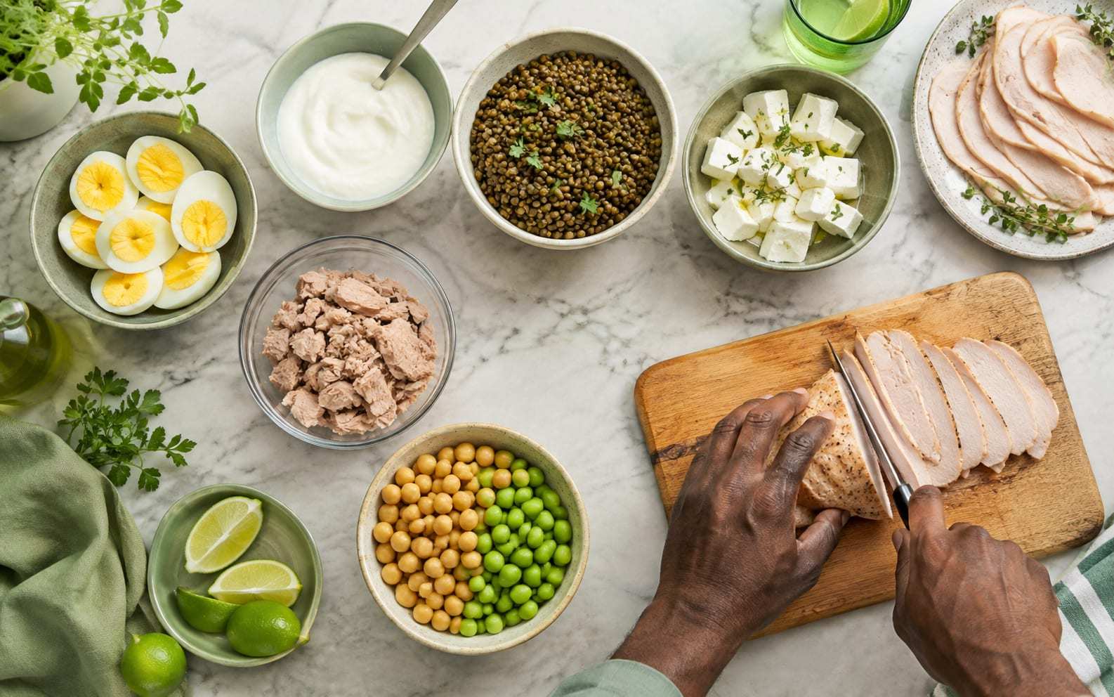 Encimera de mármol vista desde arriba con cuencos de huevos cocidos, yogur natural, lentejas, queso fresco, atún, garbanzos y guisantes, y unas manos cortando una pechuga a la plancha sobre una tabla