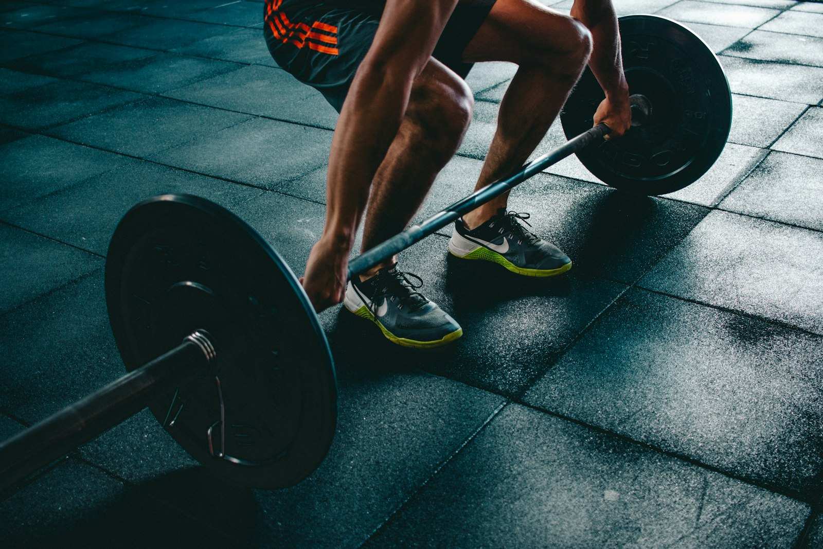 Piernas y manos de una persona en un gimnasio con suelo de caucho agarrando una barra cargada con discos negros, a punto de levantarla del suelo